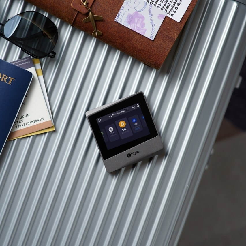 Overhead view of an ELLIPAL Titan Mini hardware wallet placed on a suitcase, surrounded by a passport and assorted travel items.