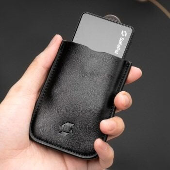 Hand holding a SafePal leather wallet with a SafePal S1 crypto hardware wallet poking out, set against a black backdrop.